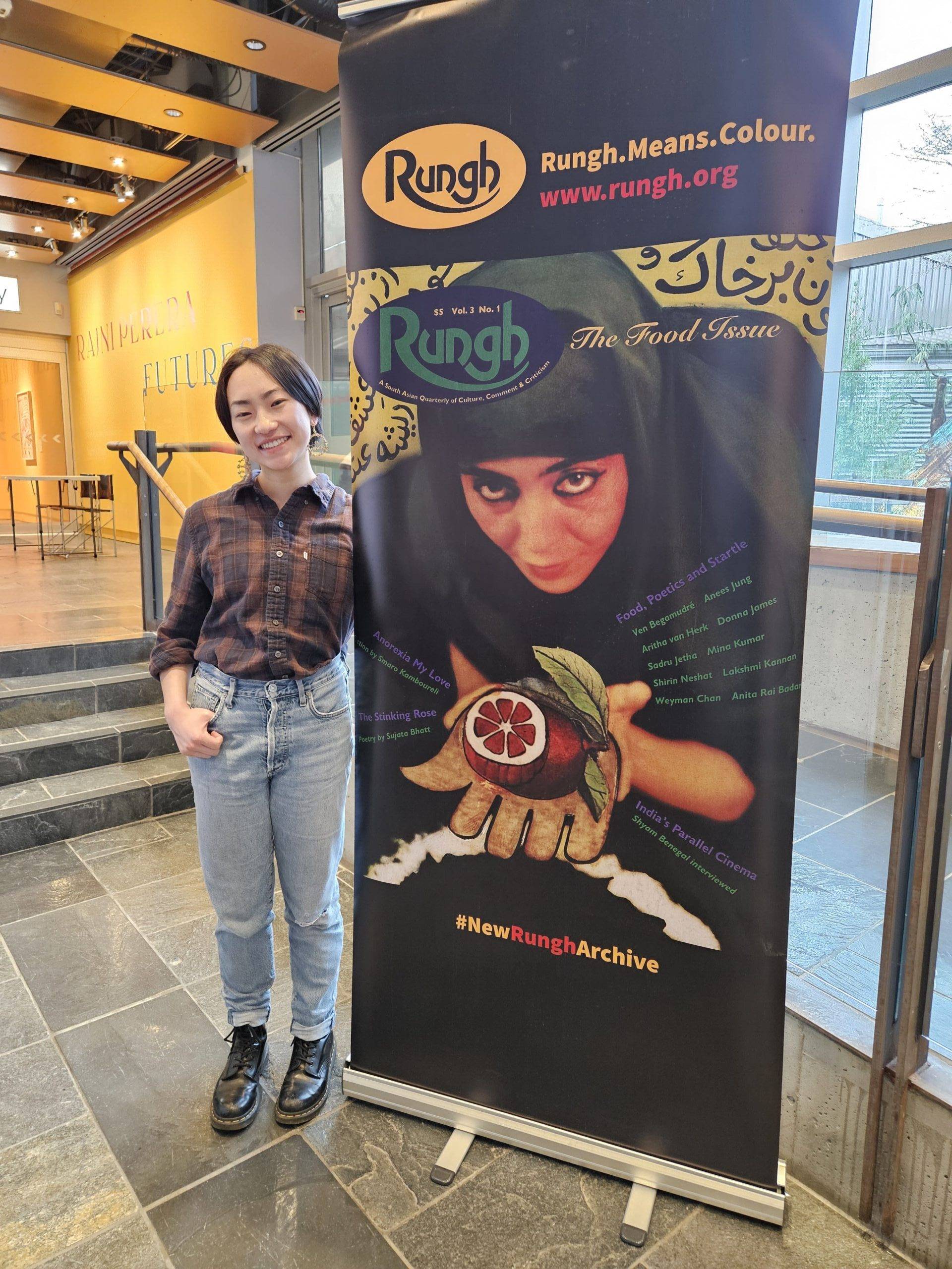 Diverse young woman at Rungh event standing beside campaign poster in a modern indoor space.