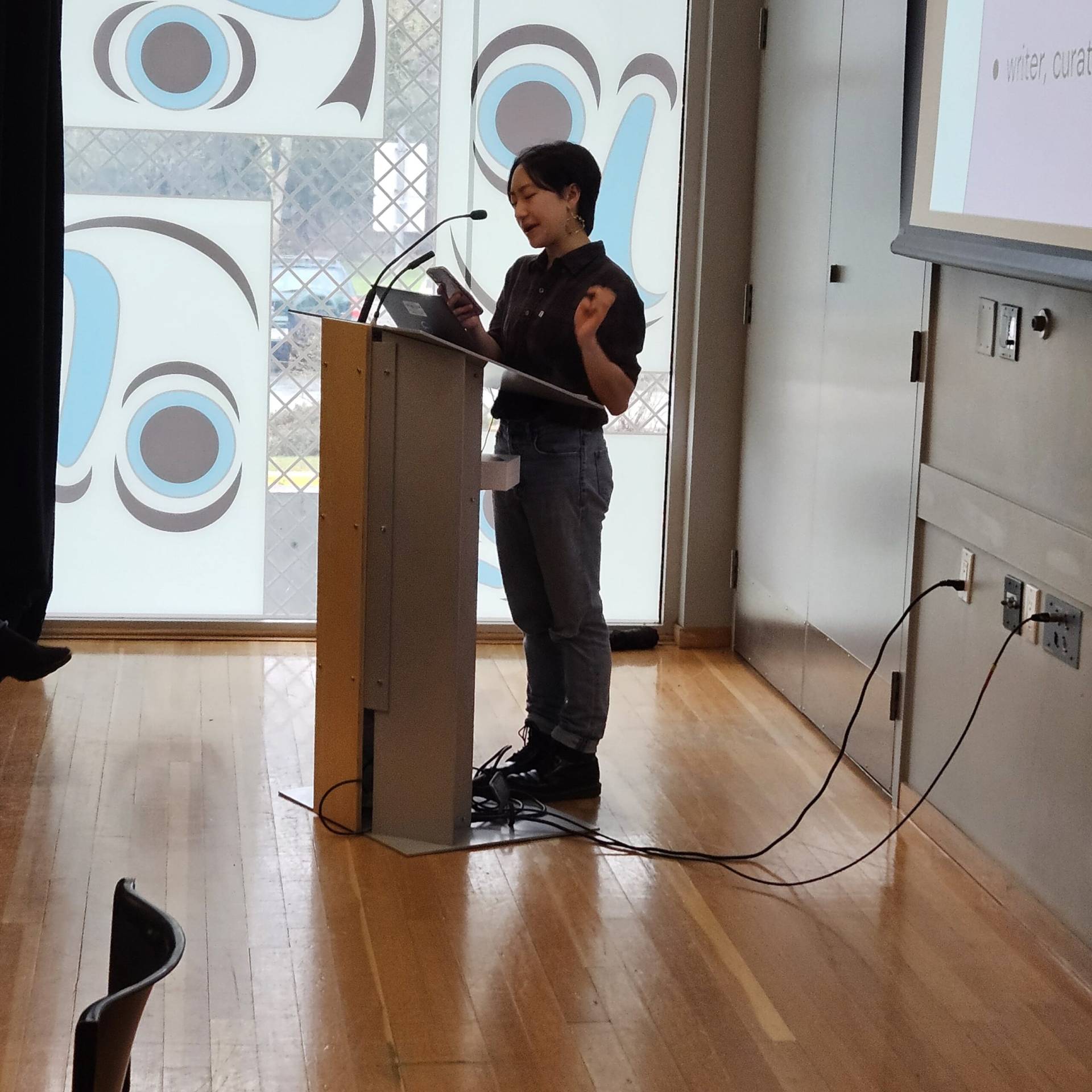 Thoughtful woman delivering a presentation at Rungh cultural centre, engaging audience on contemporary Asian arts and media.