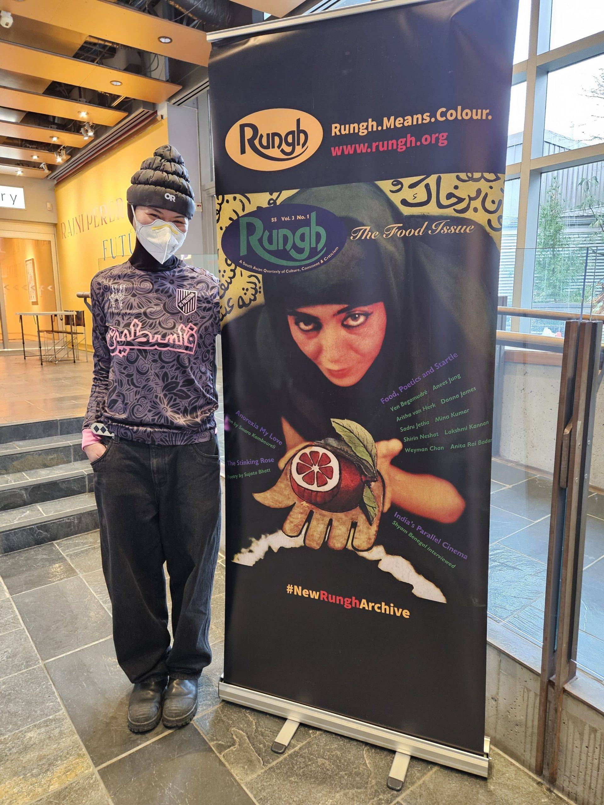 1. Diverse woman with headscarf posing beside Rungh magazine poster at cultural event.