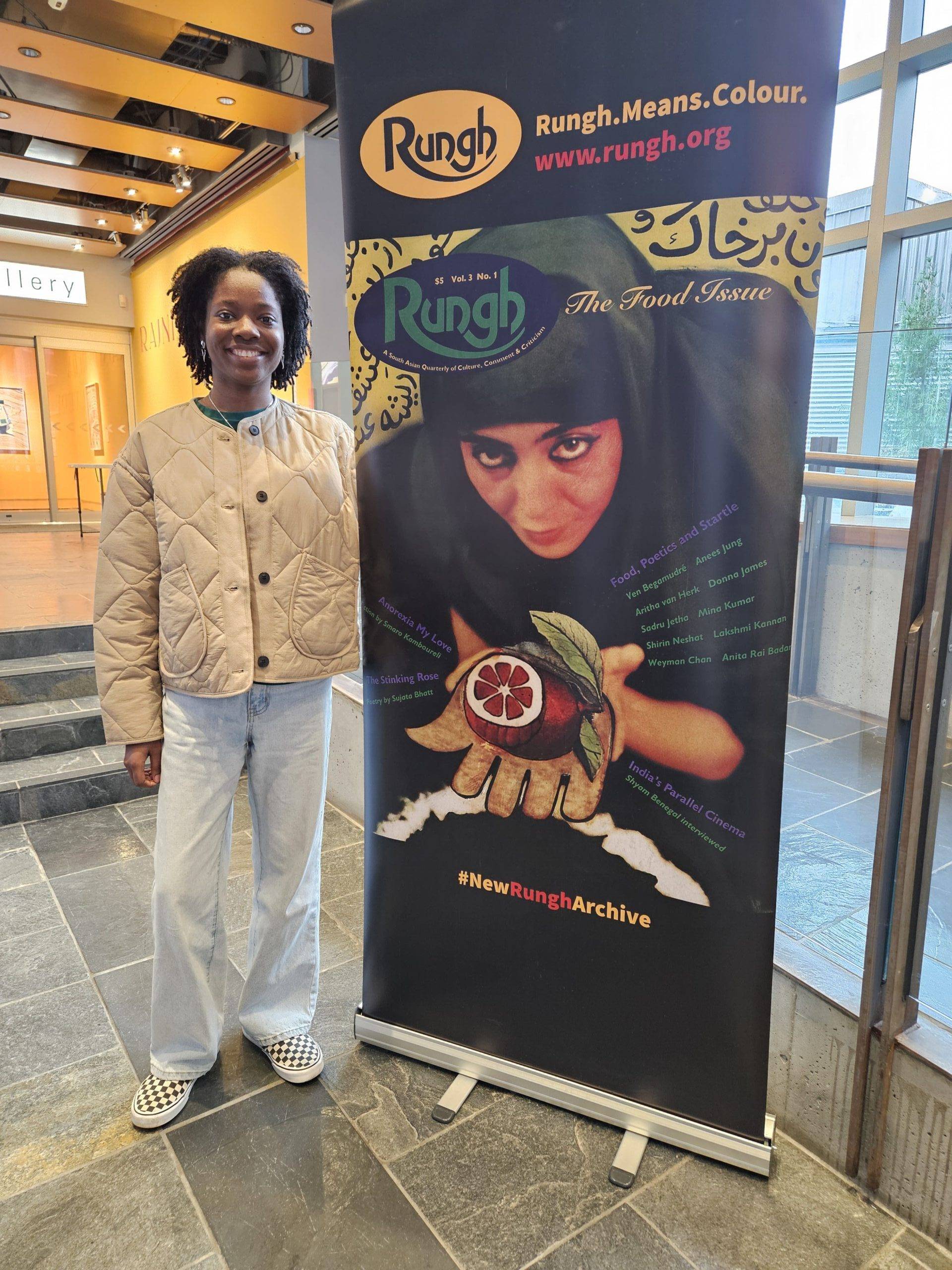 Native woman with natural hair smiling next to a Rungh magazine poster, cultural arts publication in Vancouver.