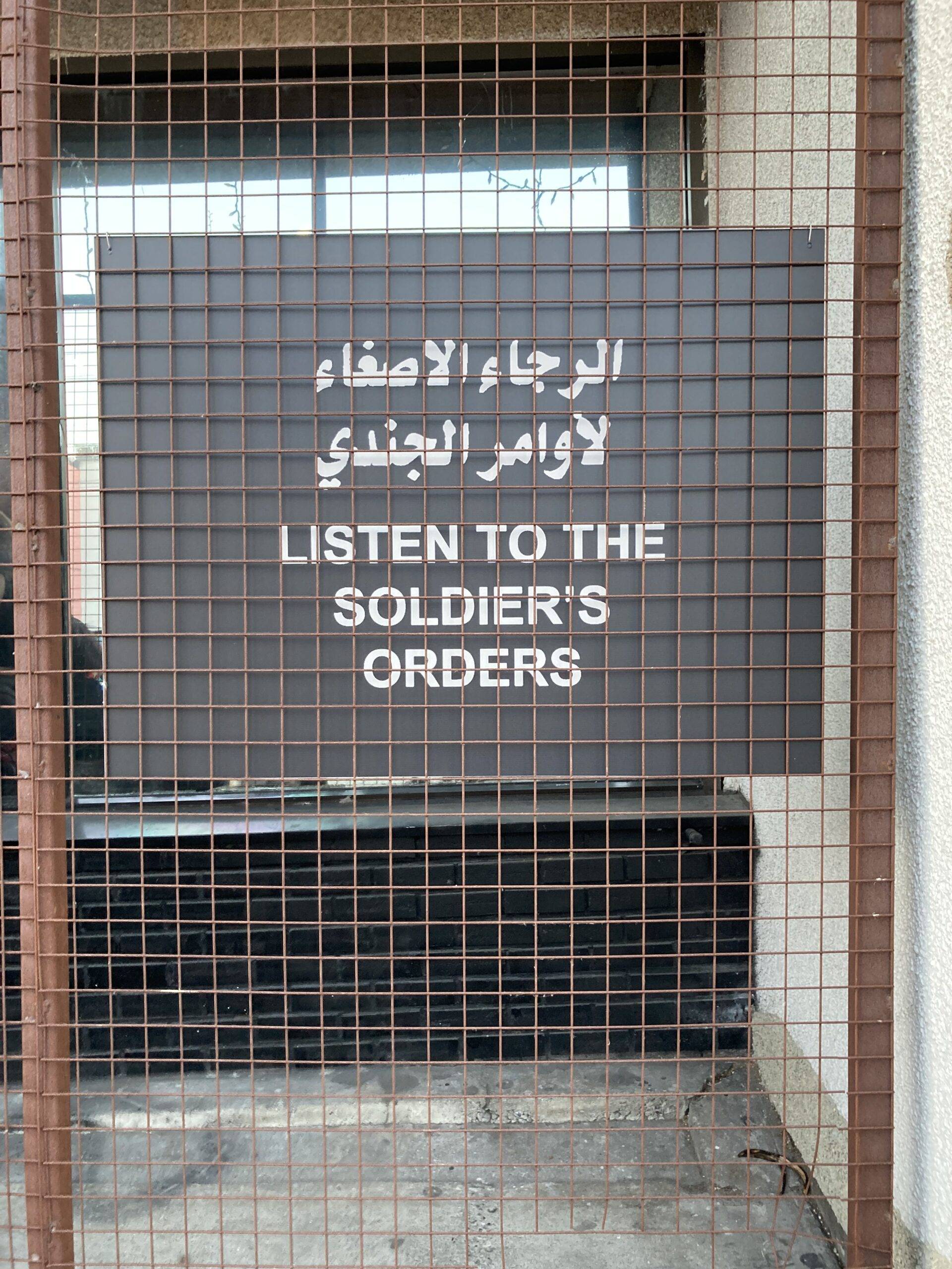 War memorial sign with Arabic and English text behind a metal grid.